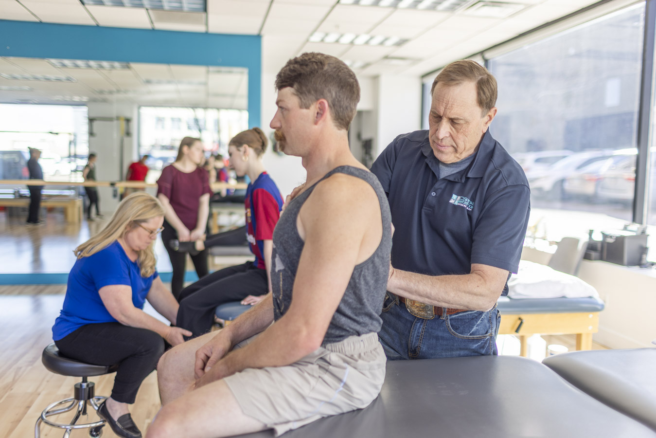 Physical therapists working with multiple patients. In the foreground, the patient is sitting facing to the left while the athletic trainer works on a spot on his back.